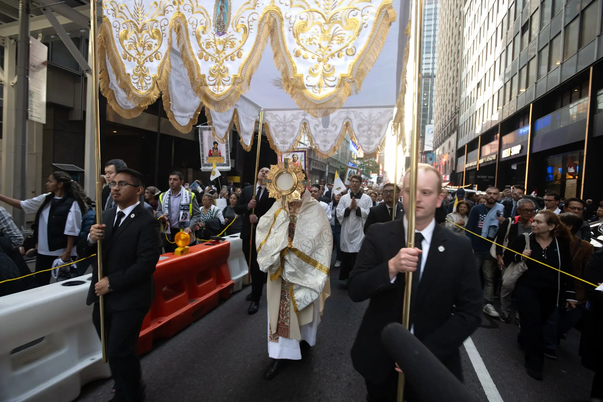 Between construction barriers and office towers, the Blessed Sacrament passes beneath a canopy of white and gold. Christ comes into the city as it is, and into our lives as they are. | Credit: Jeffrey Bruno